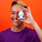 Excitedly, someone grins while holding the Cute Little Fuckers Princette Puppypus - Platinum playfully in front of their eye. The vibrant orange background highlights their purple shirt, enhancing the energetic atmosphere.